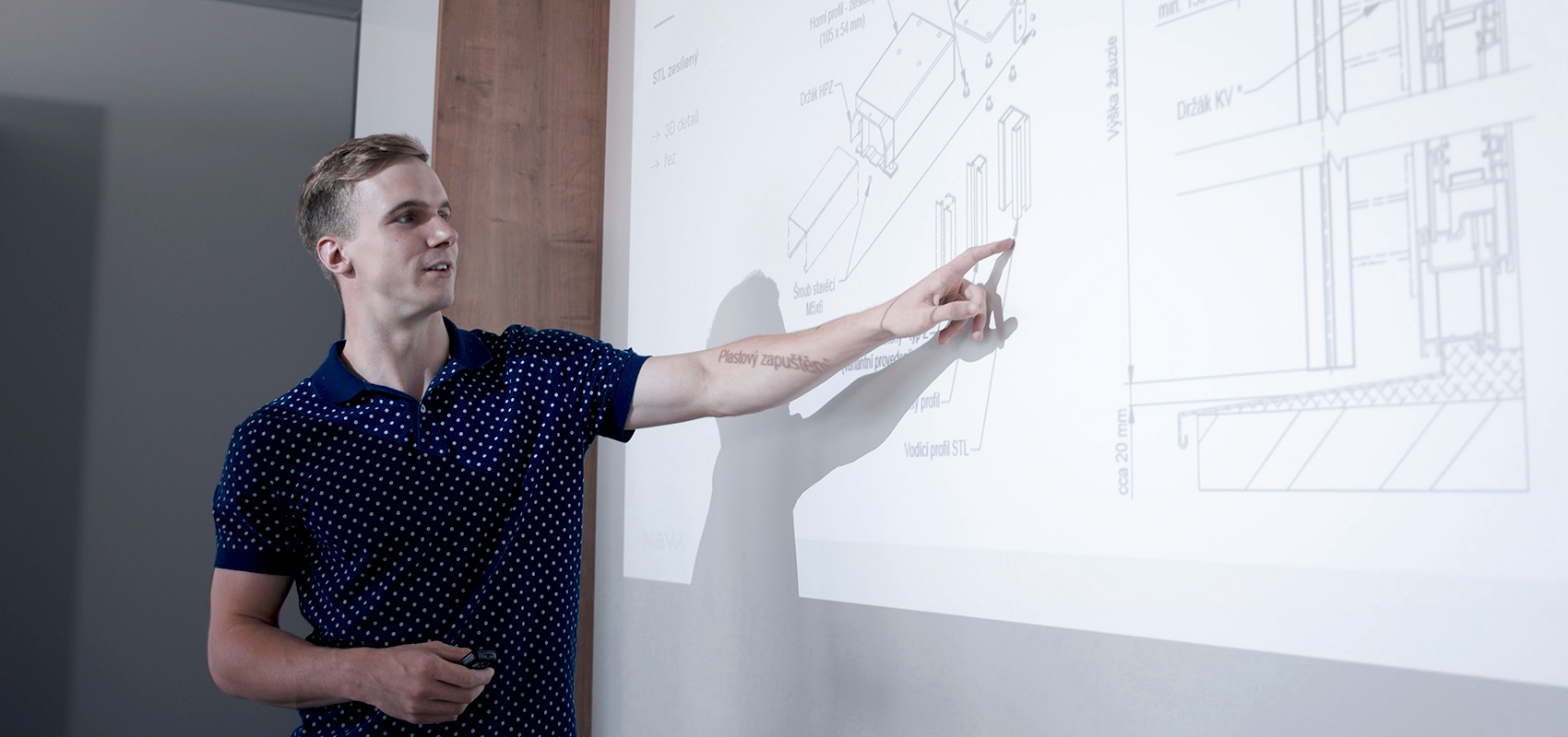 Young man in a blue polka-dot polo presenting a technical blueprint projected on a wall.